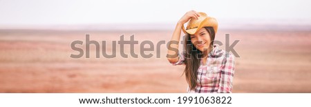 Cowgirl woman smiling happy on country farm landscape wearing cowboy hat. Beautiful young multiracial Asian American girl in panoramic countryside.