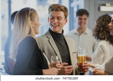 Coworkers Are Enjoying Drinks In A Bar After Work. A Man Is Holding A Pint Of Beer And Is Talking To A Woman Who Has Her Back To The Camera. 