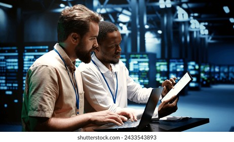 Coworkers running data center hardware diagnostic tests to determine and service software issues. Servicemen using laptop and tablet to fix critical systems, checking for blips - Powered by Shutterstock