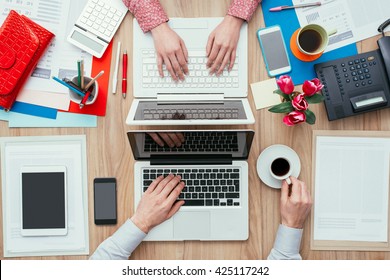 Coworkers Sharing The Same Desk With Customized Workspace And Working With A Laptop, Top View