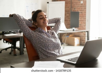 In coworking room worker indian ethnicity girl leaning on office chair put hands behind head look at pc screen read media news take break complete work relaxing. No stress end of working day concept