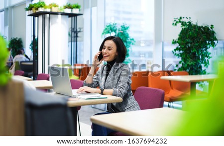 In the coworking. Side-view photo of a cheerful woman in smart casual clothes, talking on the phone while working on her laptop.