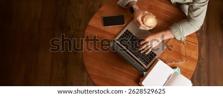 Co-working space, freelance and e-learning concept. Top view, male hands holding cup of coffee and using laptop. A man sitting in cafe and working or studying online.
