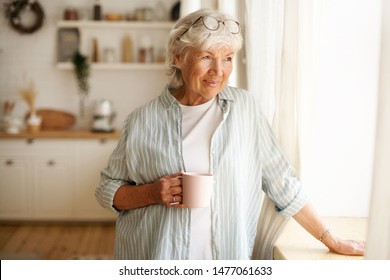 Coziness, domesticity and leisure concept. Portrait of stylish gray haired woman with round spectacles on her head enjoying morning coffee, holding mug, looking outside through window glass