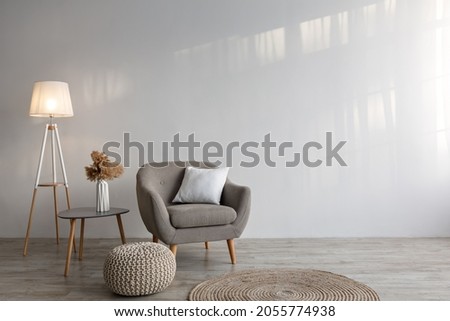 Cozy armchair with pillow, luminous lamp, table, dry plants in vase, ottoman and carpet on floor on gray wall background in living room. Boho style, modern design and blog about interior and furniture