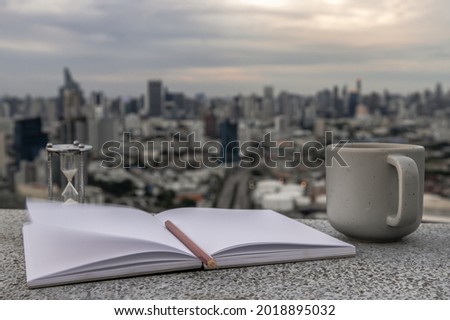 Cozy atmosphere with Evening coffee with a city view. There is a cup of coffee, hourglass, diaries, notebook, pencil to record memories, Workplace area and blurry urban view, Selective focus.