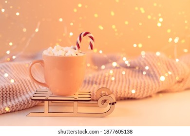 A Cozy Atmosphere Of Home Evening Relaxation, A Delicate Pastel Pink Blanket, A Pink Mug Of Cocoa With Marshmallows And Christmas Caramel Canes In The Glow Of A New Year Garland, Sleigh. Front View.