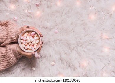 Cozy Background. Hands Holding Mug Of Hot Cocoa Or Hot Chocolate With Marshmallow On White Rug. Copy Space