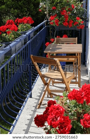 A cozy balcony offers a vibrant display of red geraniums beside a charming wooden table and chairs.