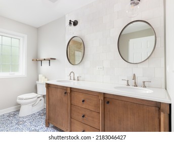 A Cozy Bathroom With A Patterned Tile Floor, Natural Wood Vanity, Tiled Backsplash, And Lights Mounted Above Circular Mirrors.