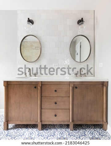 A cozy bathroom with a patterned tile floor, natural wood vanity, tiled backsplash, and lights mounted above circular mirrors.