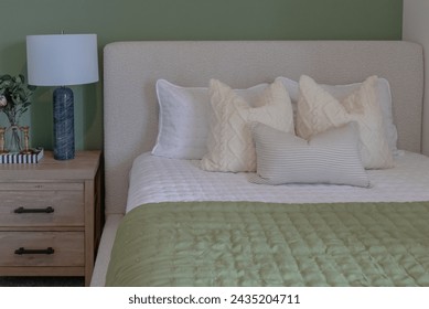 Cozy bed with green comforter or duvet with many white pillows, upholstered headboard and wood side table