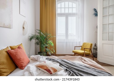 Cozy bedroom in light apartment with interior design in the style of the boho chic. Comfortable bed with colorful cushions against potted houseplant and bright yellow armchair in white room