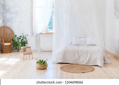Cozy bedroom in light colors with a wooden floor, a large four-poster bed, wicker chair and basket of flowers. Scandinavian simplicity design. Eco loft apartments.