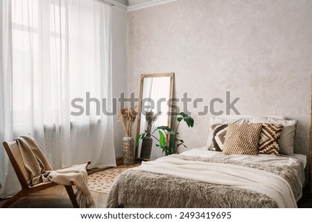 Cozy bedroom with plaid and decorative cushions on large bed, soft blanket on wooden chair and mirror close to window white tulle or curtains. Interior design in boho style apartment