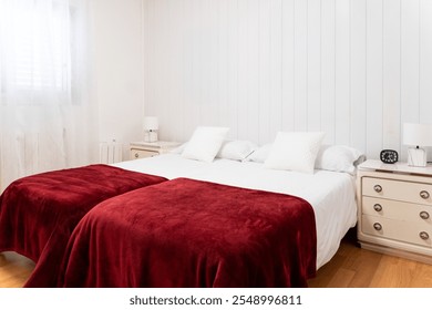 Cozy bedroom with white walls, wooden floor, and bedside tables featuring two single beds with red blankets - Powered by Shutterstock