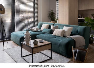 Cozy, blue corner sofa with square, wooden coffee table with decorations in modern living room with big windows with blinds