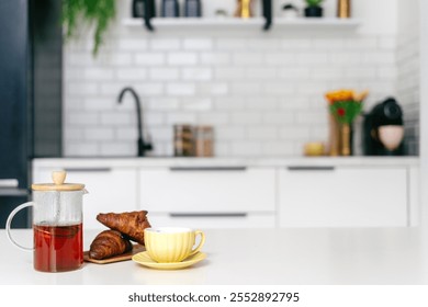 A cozy breakfast is arranged with a glass teapot, buttered croissants and a yellow cup in a stylish kitchen setting. French press with hot tea close to fresh pastry - Powered by Shutterstock