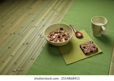 A cozy breakfast with a bowl of granola, dried fruits, white chocolate chips, a wooden spoon, granola bar, and cup of milk, all arranged on a green placemat and bamboo mat beneath. - Powered by Shutterstock