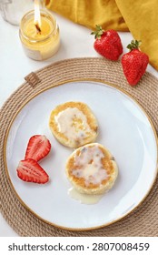 A cozy breakfast flat lay featuring two golden mini cheesecakes drizzled with creamy glaze on a white plate. Served with fresh strawberries, next to a burning candle and mustard yellow fabric on a wov
