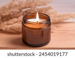 Cozy burning candle in amber glass jar, winter hygge home interior decor on wooden tray with dry reeds or pampas grass close up. Scented wax eco candle.