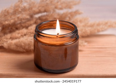 Cozy burning candle in amber glass jar, winter hygge home interior decor on wooden tray with dry reeds or pampas grass close up. Scented wax eco candle. - Powered by Shutterstock