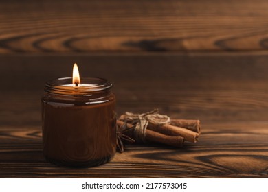 A cozy burning candle in a brown glass jar with cinnamon sticks and pumpkin candles on a brown wood texture. winter home decor. Soy ecological candle. Home and interior decoration. Place for text. Cop - Shutterstock ID 2177573045