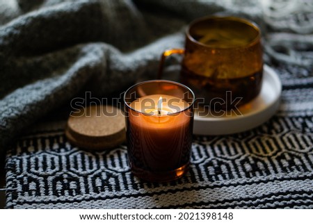 Cozy burning candle in brown glass jar, winter home interior decor with cup and blanket