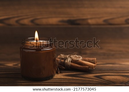 A cozy burning candle in a brown glass jar with cinnamon sticks and pumpkin candles on a brown wood texture. winter home decor. Soy ecological candle. Home and interior decoration. Place for text. Cop