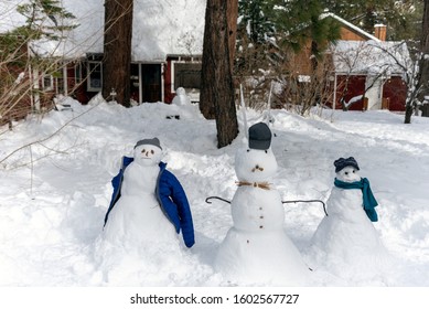 Cozy Cabin With Pine Trees, Roof, And Yard Covered In Fresh Snow With Three Members Of A Snowman Family Dressed In Winter Clothes In Wrightwood, California.