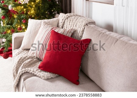 Cozy Christmas Ambiance, A Neutral-Toned Sofa Adorned with Textured Cushions, Rich Red Accent Pillow, Chunky Knit Throw, Set Against a Glittering Christmas Tree Illuminated by Golden Lights, Close Up