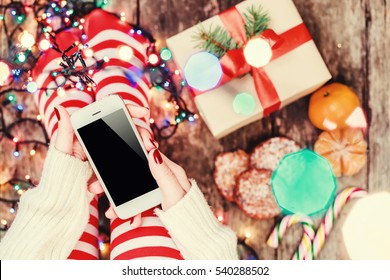 Cozy Christmas at home. Female hands holding mobile phone. Women's feet in Christmas stockings, big present on bokeh light background. Top view. Selective focus. Toning - Powered by Shutterstock