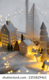 Cozy christmas miniature village. Stylish little ceramic houses and wooden trees on soft snow blanket with glowing lights in evening. Atmospheric winter village still life. Merry Christmas!
