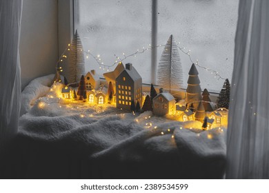 Cozy christmas miniature village. Stylish little ceramic houses and wooden trees on soft snow blanket with glowing lights in evening room. Atmospheric winter village still life. Merry Christmas! - Powered by Shutterstock