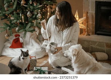 Cozy christmas morning. Beautiful woman in pajamas opening stylish christmas gift with cute white dog and cat at fireplace and decorated tree in festive living room. Merry Christmas! - Powered by Shutterstock