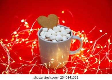 A cozy Christmas scene: a cup of hot cocoa with marshmallows and a heart-shaped gingerbread cookie on a red background, illuminated by warm string lights for a festive, holiday atmosphere. - Powered by Shutterstock