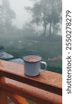 A cozy coffee cup sits on a wooden railing during a misty, rainy day. The serene scene captures a peaceful moment overlooking a lush forest backdrop