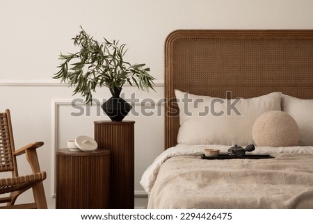 Cozy composition of bedroom interior with cozy bed, beige bedding, wooden bedside table, rattan armchair, patterned pillow and personal accessories. Home decor. Template. 