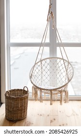 Cozy corner at home with hanging armchair macrame near the window. Simple interior in eco-style: wooden floor, macrame swing, straw wicker basket. A place of rest and relaxation