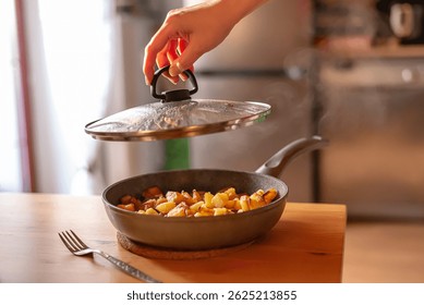 Un momento culinario acogedor: una mano levanta la tapa de una sartén sobre una mesa de madera, revelando una deliciosa comida. Una iluminación suave, una ventana con cortinas y flores en el exterior crean un ambiente cálido y hogareño