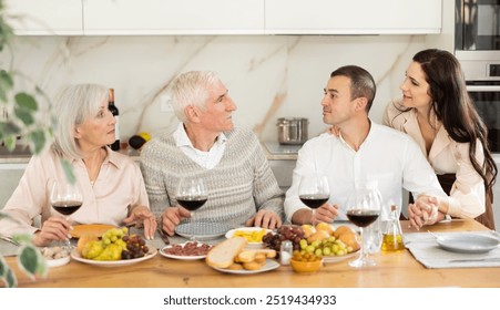 Cozy dinner scene while elderly parents enjoying wine and meal with adult daughter and son-in-law in well-lit home kitchen..