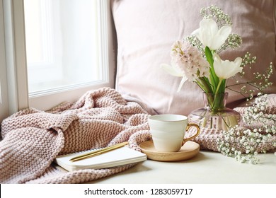 Cozy Easter, spring still life scene. Cup of coffee, opened notebook, pink knitted plaid on windowsill. Vintage feminine styled photo. Floral composition with tulips, hyacinth and Gypsophila flowers.