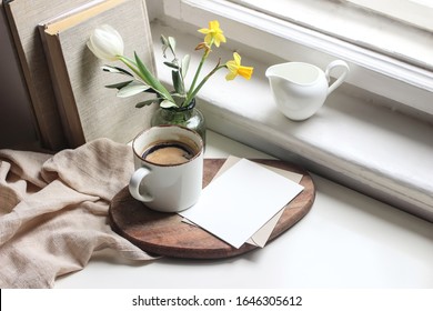 Cozy Easter spring still life. Greeting card mockup scene. Cup of coffee, books, wooden cutting board, milk pitcher and vase of flowers on windowsill. Floral composition. Yellow daffodils and tulip.  - Powered by Shutterstock