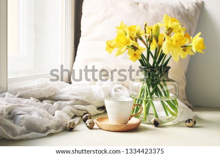 Cozy Easter, spring still life scene. Cup of coffee, wooden plate, quail eggs and vase of flowers on windowsill. Floral composition with yellow daffodils, narcissus. Vintage feminine styled photo. 