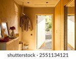 A cozy entryway featuring hanging clothes, a wooden console with a flower vase, and an open door letting in warm sunlight, revealing the natural outdoor landscape