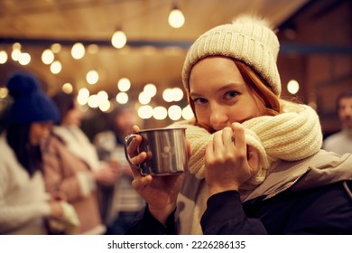 Cozy evening. Close-up of young beautiful girl in winter attire drinking hot drinks and dreaming, making a wish. Spending time with friends at winter fair at evening. Looks happy, delighted. - Shutterstock ID 2226286135