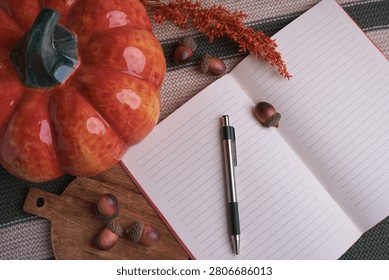 Cozy fall flat lay: open lined notebook with pen, orange ceramic pumpkin, and acorns on a wooden board over knitted textiles.