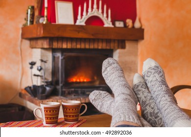 Cozy Family Evening At Home Near Fireplace In Winter, Feet Of Couple In Socks And Two Cups Of Tea, Weekend