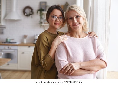 Cozy Family Portrait Of Stylish Cute Teenage Girl In Eyeglasses Embracing Her Charming Beautiful Middle Aged Mother. Attractive Neat Blonde Female Posing In Kitchen With Her Young Daughter