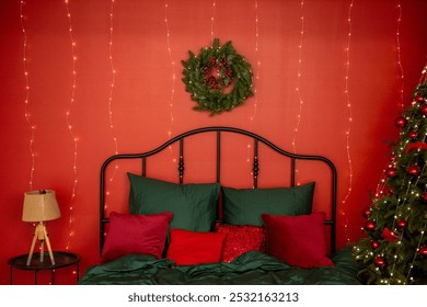 Cozy, festive bedroom decorated for holidays. Red wall adorned with string lights, green bedding set, Christmas wreath. Christmas tree with red ornaments stands in the corner. Copy space - Powered by Shutterstock